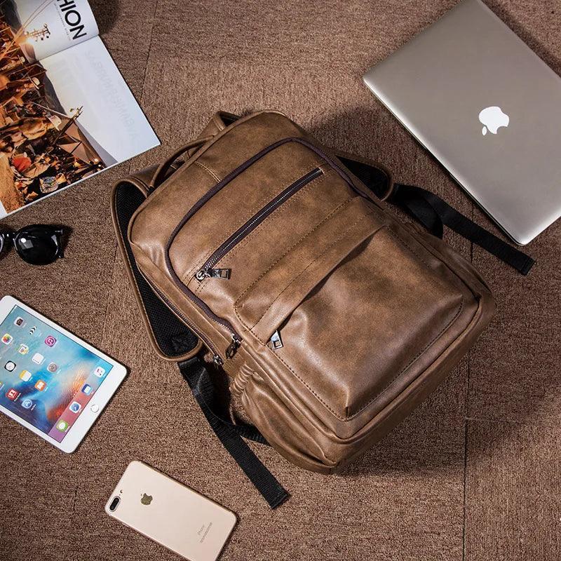 Mochila masculina de cuero marrón compacta sobre alfombra, junto a laptop, tablet y revista.